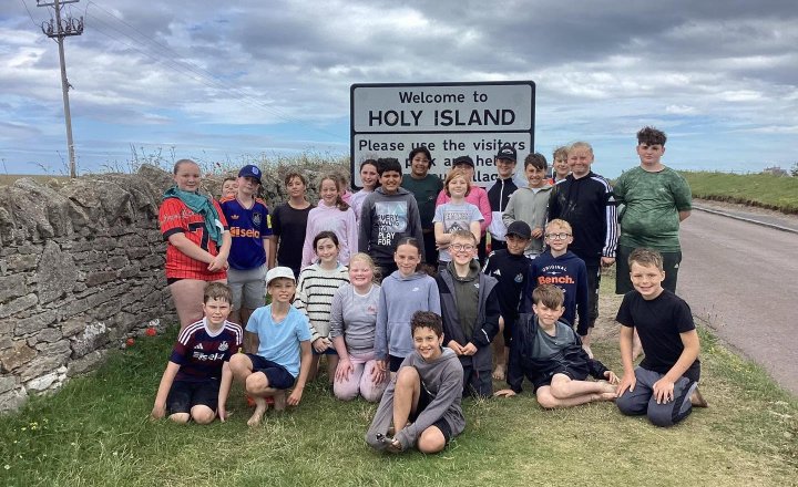 Year 6 pupils from Holy Family Primary School in Darlington stand and kneel together on grass in front of a “Welcome to Holy Island” sign
