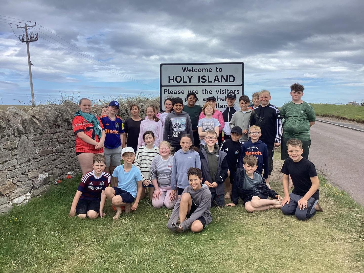 Year 6 pupils from Holy Family Primary School in Darlington stand and kneel together on grass in front of a “Welcome to Holy Island” sign
