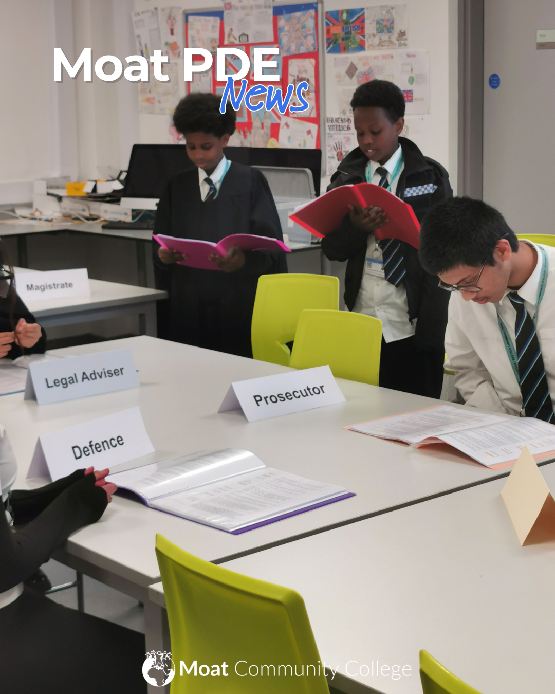 An image of three students in a classroom. The first student is wearing a black robe, the second a police officer’s uniform. The third student is seated at a table and is studying some paperwork. 