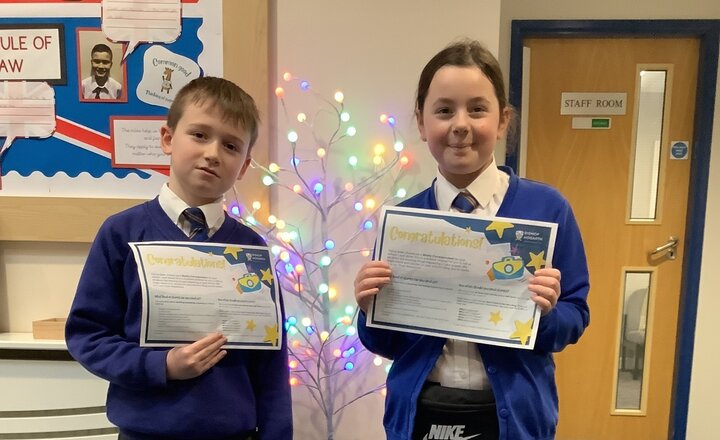 Two Year Five pupils in school uniform proudly holding certificates while standing in front of a decorative tree with colourful lights.