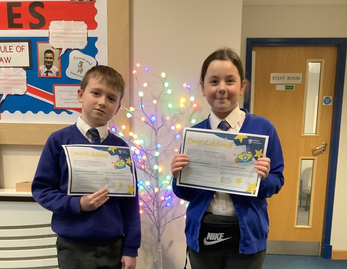Two Year Five pupils in school uniform proudly holding certificates while standing in front of a decorative tree with colourful lights.