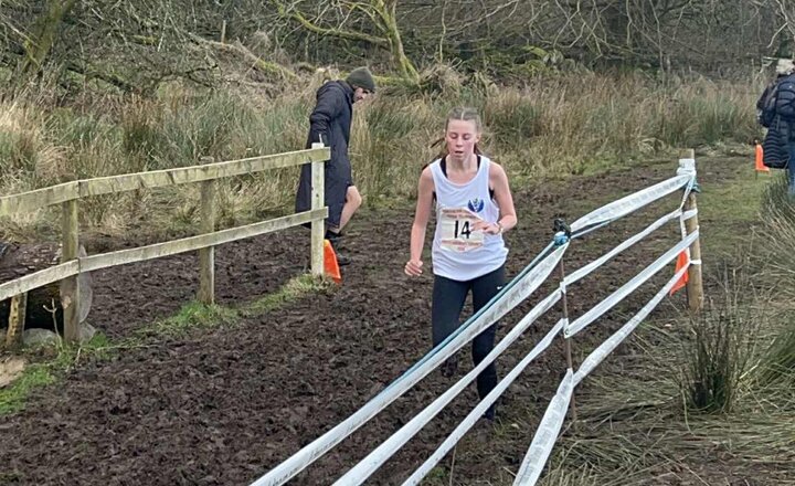 A young cross‑country runner wearing a competition vest runs along a muddy outdoor course lined with tape, with trees and spectators in the background.