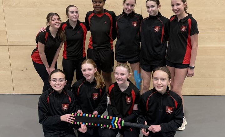  A group of U13 girls’ cricket players wearing matching black and red sports kits pose together indoors, with two pupils at the front holding a colourful cricket bat.