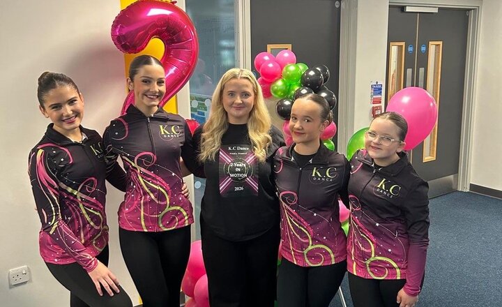 A group of students wearing matching KC Dance performance tops stand together indoors with their dance coach, surrounded by colourful balloons.