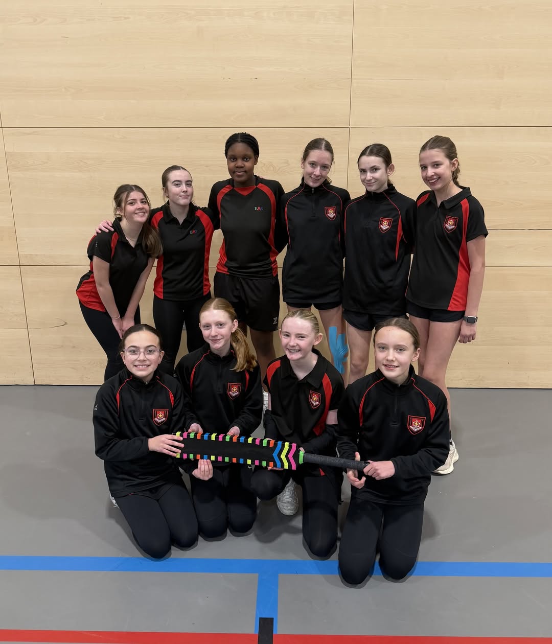 A group of U13 girls’ cricket players wearing matching black and red sports kits pose together indoors, with two pupils at the front holding a colourful cricket bat.